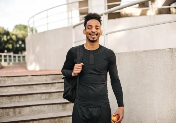 Homem jovem sorrindo ao ar livre, com mochila e uma maçã na mão